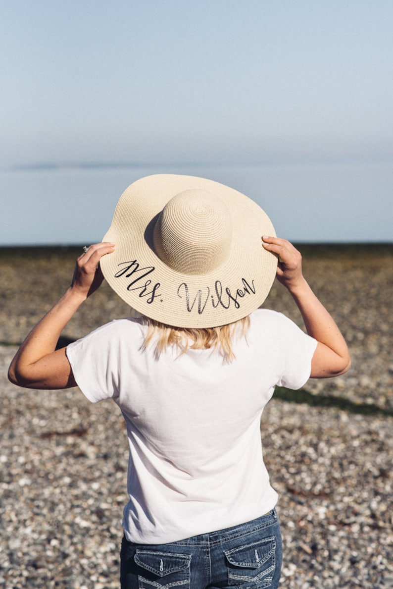 Puede incluir: Una mujer con una camiseta blanca y pantalones vaqueros azules lleva un sombrero de paja para el sol con el texto "Mrs. Wilson" escrito en &eacute;l.