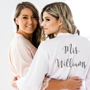 May include: Two women wearing white and pink satin robes with lace trim. The woman in the white robe has the text "Mrs. Williams" printed on the back.