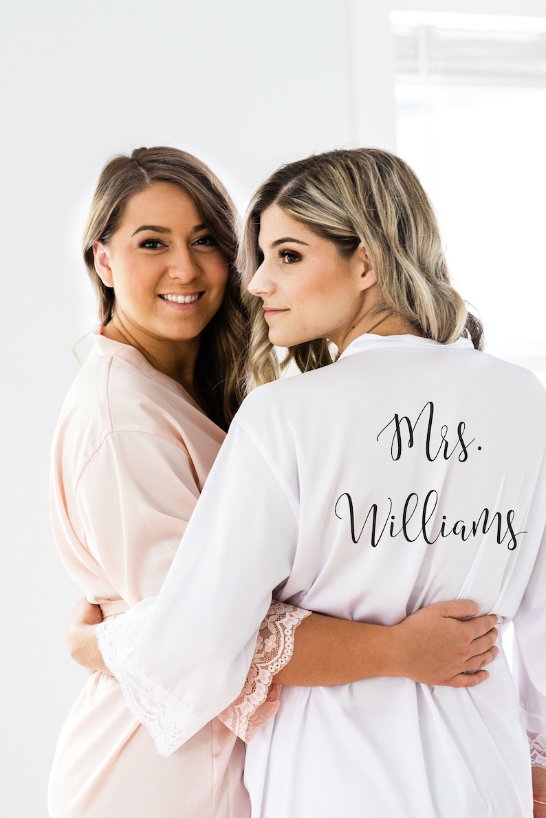 May include: Two women wearing white and pink satin robes with lace trim. The woman in the white robe has the text "Mrs. Williams" printed on the back.