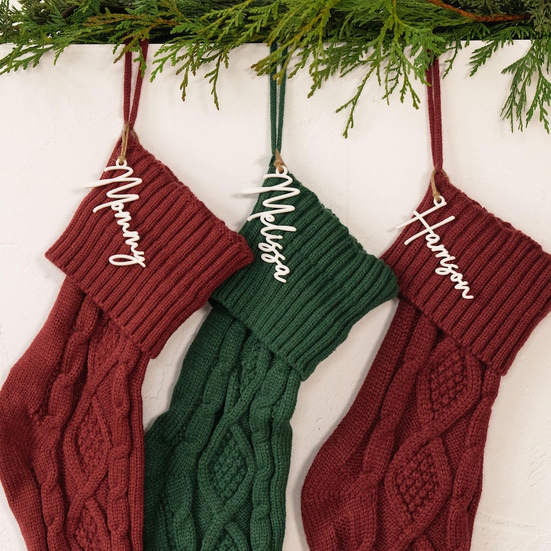 May include: Three cable-knit Christmas stockings in burgundy and green hang from a branch. Each stocking has a white name tag: "Mommy", "Melissa", and "Harrison". The stockings are suspended by twine.