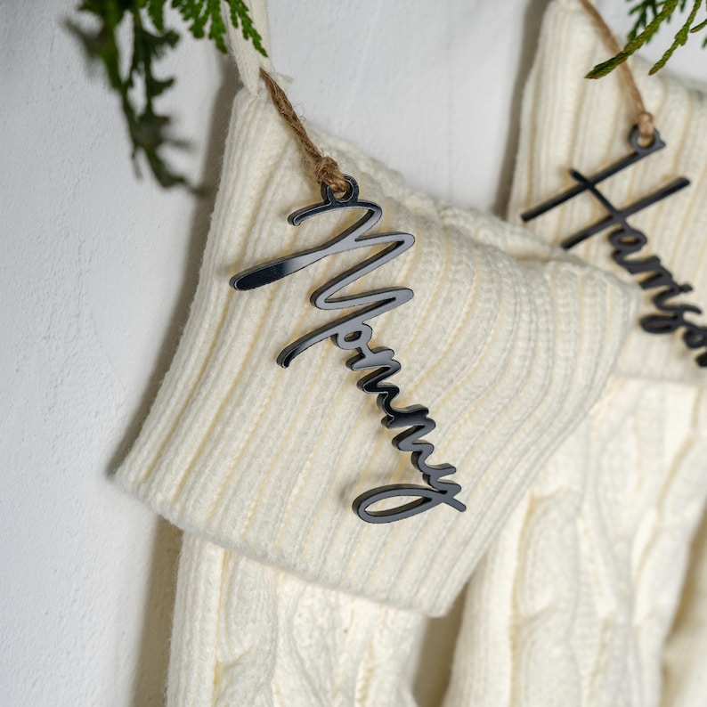 May include: Close-up of a cream-colored Christmas stocking with a silver-colored "Mommy" name tag hanging from a twine string. The stocking has a cable knit design. A second stocking with a name tag is partially visible.
