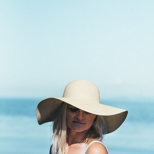 Puede incluir: Una mujer con un sombrero de sol blanco de ala ancha, un top blanco sin mangas y cabello rubio. El fondo es un cielo azul y agua.