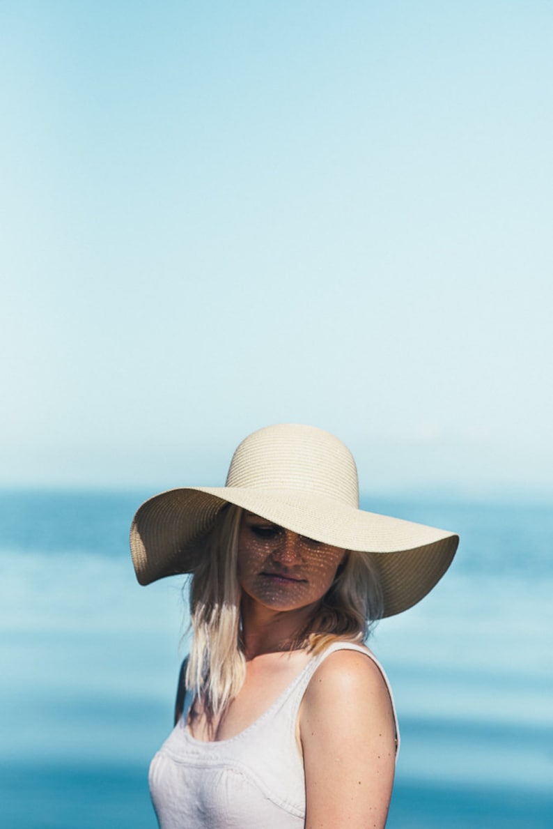 Puede incluir: Una mujer con un sombrero de sol blanco de ala ancha, un top blanco sin mangas y cabello rubio. El fondo es un cielo azul y agua.