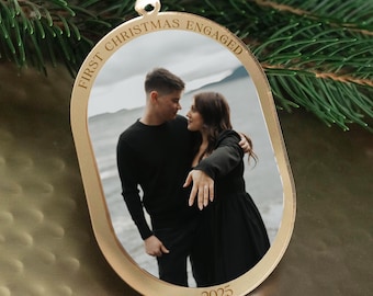 Op de afbeelding: Goudkleurige kerstbal met een foto van een stel. De ornament is gegraveerd met "FIRST CHRISTMAS ENGAGED" en het jaar "2025". Een wit lint is bevestigd om op te hangen.