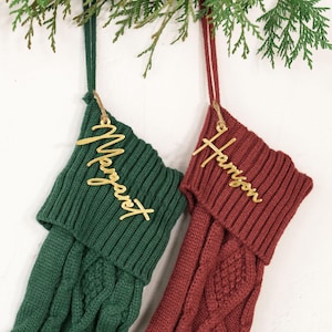 May include: Two Christmas stockings, one green and one burgundy, hang from a branch. Each stocking has a gold name tag. The green stocking has the name "Margaret" and the burgundy stocking has the name "Harrison".