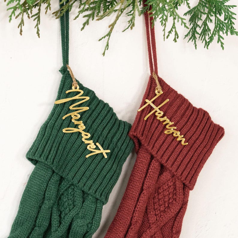 May include: Two Christmas stockings, one green and one burgundy, hang from a branch. Each stocking has a gold name tag. The green stocking has the name "Margaret" and the burgundy stocking has the name "Harrison".