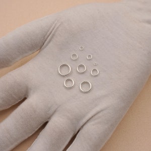 May include: Assortment of silver jump rings in various sizes, displayed on a white surface.