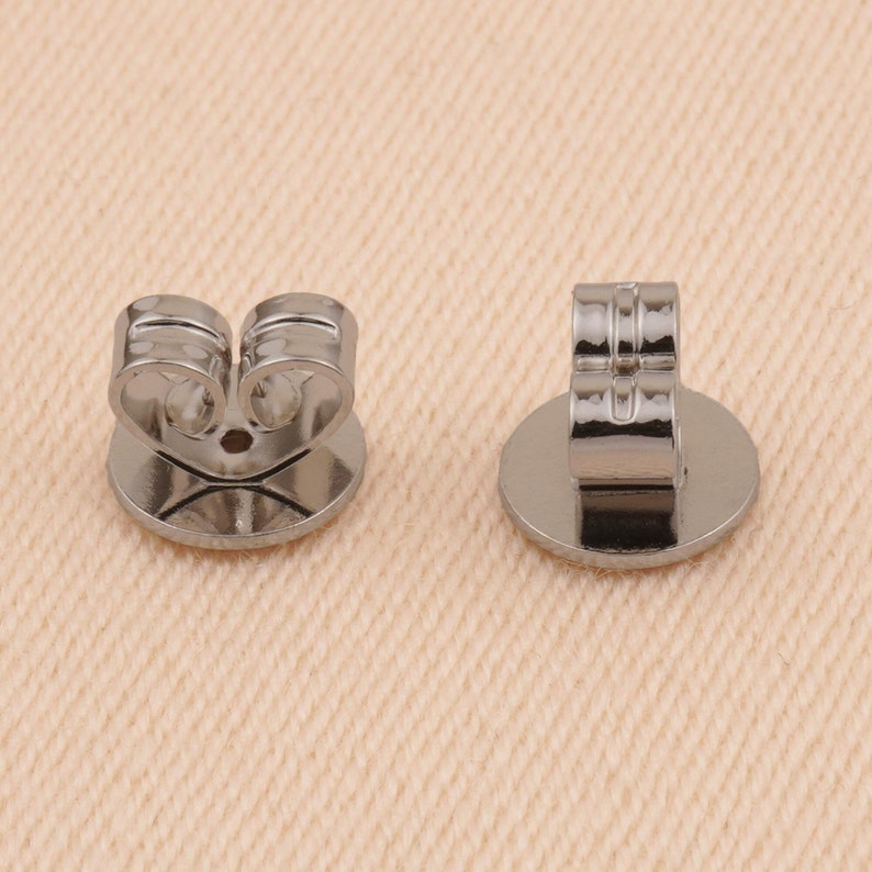 May include: Two silver earring backs, one with the back facing the camera and the other with the front facing the camera.