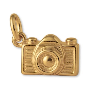 Puede incluir: Un dije dorado con forma de cámara. El dije tiene un lazo en la parte superior para sujetarlo a un collar o pulsera. La cámara tiene un diseño detallado, que incluye una lente, botones y un cuerpo texturizado.