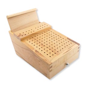 May include: A light-colored wooden cribbage board featuring a rectangular playing surface with numerous drilled holes arranged in rows. The board includes a hinged lid that opens to reveal a storage compartment and a small silver-toned metal knob.