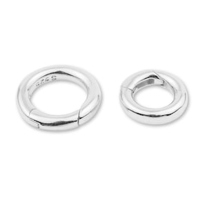May include: Two silver-coloured, round clasps. The larger clasp has the text "S 925" engraved on its surface. Both clasps are open, revealing their hinged design. The clasps are likely used for jewellery making or as closures for accessories.