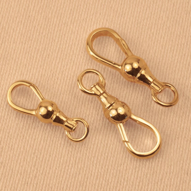 May include: Three gold-toned metal swivel lobster claw clasps, each featuring a smooth, rounded central ball joint, a teardrop-shaped opening mechanism, and a small circular attachment ring at the base for jewelry making or accessory hardware.