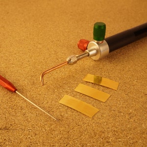 May include: A close-up of a soldering torch with a copper tip and a black handle. Three gold-colored metal sheets are laid out on a brown cork surface. A red and silver soldering tool is also visible.