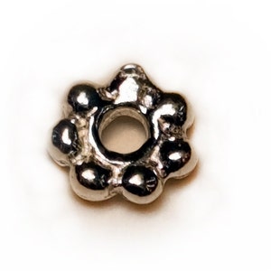 Könnte beinhalten: Silberfarbener Metallperle mit einem blumenförmigen Design und einem runden Loch in der Mitte.
