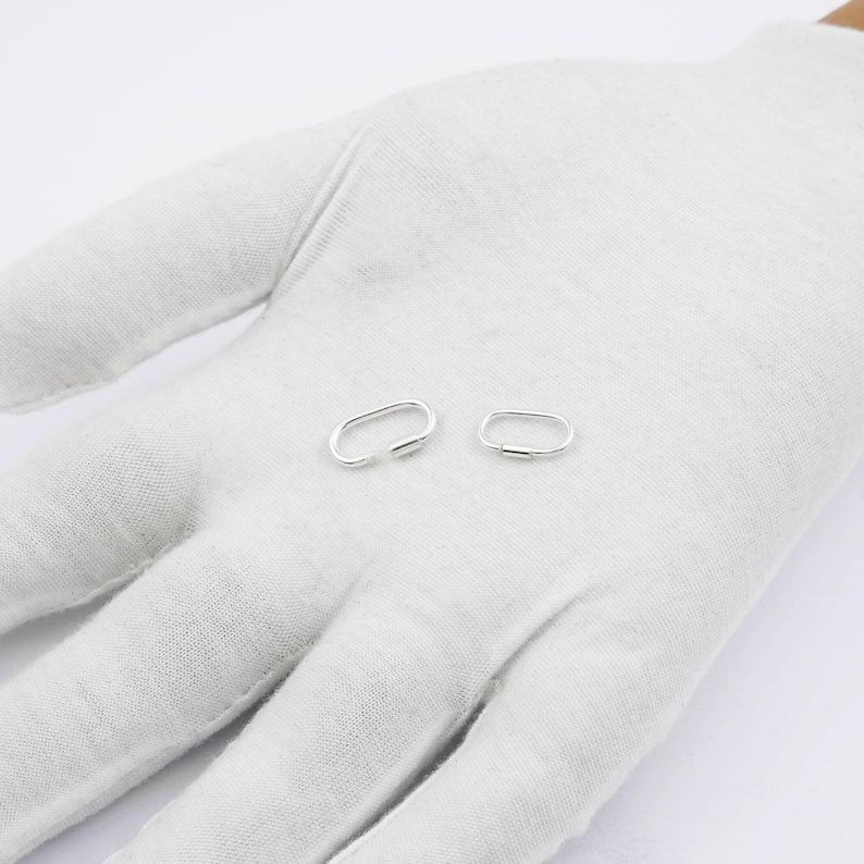 K&ouml;nnte beinhalten: Zwei kleine, ovale Ohrringe aus Silber. Ein Ohrring ist offen, der andere geschlossen. Die Ohrringe werden auf einer wei&szlig;en Oberfl&auml;che, wahrscheinlich einer behandschuhten Hand, vor einem schlichten wei&szlig;en Hintergrund pr&auml;sentiert. Die Ohrringe sind schlicht und minimalistisch.