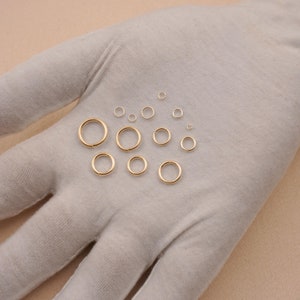 May include: A set of 12 gold jump rings of varying sizes, displayed on a white fabric surface.