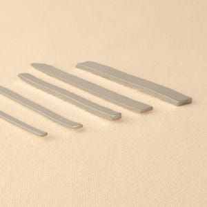 May include: Five silver metal rods of varying lengths, arranged in descending order of size, on a beige surface.