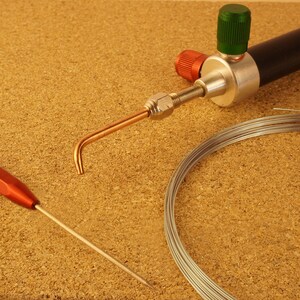 May include: A close-up of a silver soldering torch with a green and red knob, a copper tip, and a coil of silver wire.