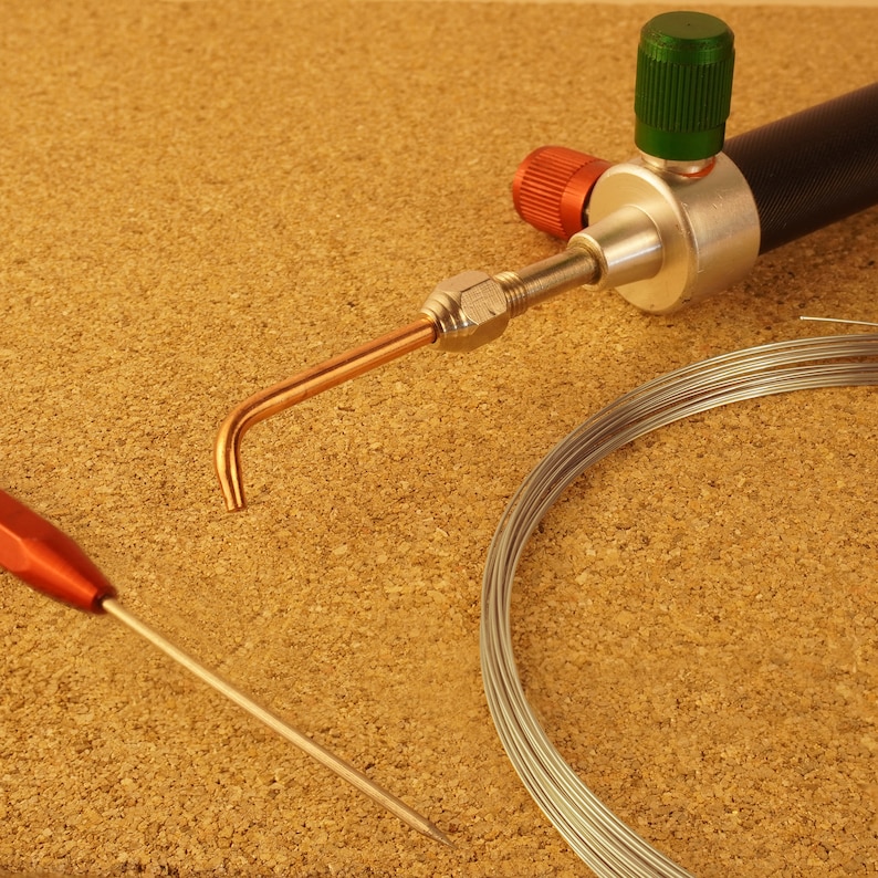 May include: A close-up of a silver soldering torch with a green and red knob, a copper tip, and a coil of silver wire.