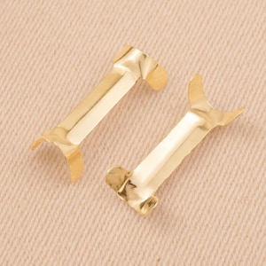 Puede incluir: Dos fornituras met&aacute;licas de color dorado. Cada fornitura tiene un cuerpo rectangular con extremos curvos. Las fornituras est&aacute;n dise&ntilde;adas para la fabricaci&oacute;n de joyas u otros proyectos de artesan&iacute;a. Se muestran sobre una superficie de color beige claro.