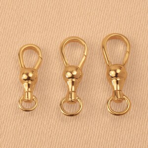 May include: Three gold-toned metal swivel lobster clasps arranged in a row from smallest to largest. Each clasp features a smooth, rounded bulbous center, a teardrop-shaped top loop, and a small circular ring attached to the bottom base.
