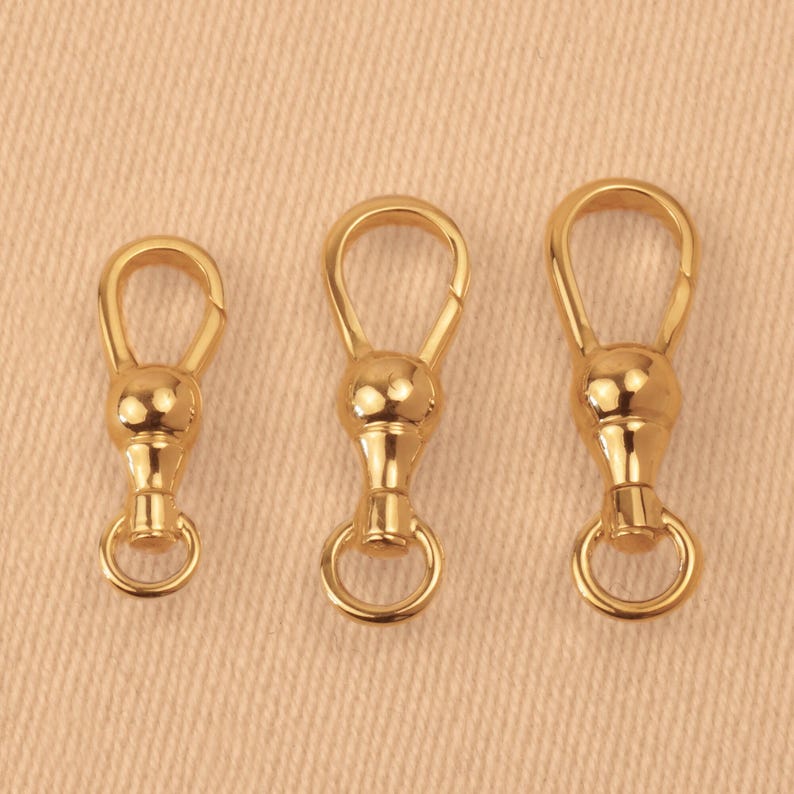 May include: Three gold-toned metal swivel lobster clasps arranged in a row from smallest to largest. Each clasp features a smooth, rounded bulbous center, a teardrop-shaped top loop, and a small circular ring attached to the bottom base.