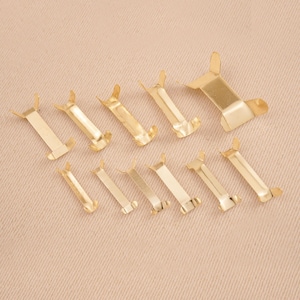 Puede incluir: Una colecci&oacute;n de diez peque&ntilde;os clips met&aacute;licos dorados dispuestos sobre una superficie beige. Cada clip tiene un cuerpo rectangular con dos extensiones curvas en forma de ala en cada extremo. Los clips var&iacute;an ligeramente en tama&ntilde;o.