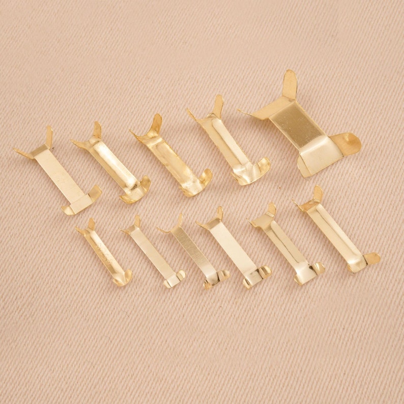 Puede incluir: Una colecci&oacute;n de diez peque&ntilde;os clips met&aacute;licos dorados dispuestos sobre una superficie beige. Cada clip tiene un cuerpo rectangular con dos extensiones curvas en forma de ala en cada extremo. Los clips var&iacute;an ligeramente en tama&ntilde;o.