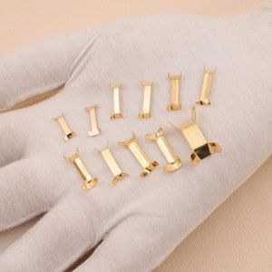 Puede incluir: Una colecci&oacute;n de peque&ntilde;os cierres met&aacute;licos dorados y brillantes. Los cierres son rectangulares con un dise&ntilde;o curvo en forma de ala en cada extremo. Est&aacute;n dispuestos sobre una superficie blanca.