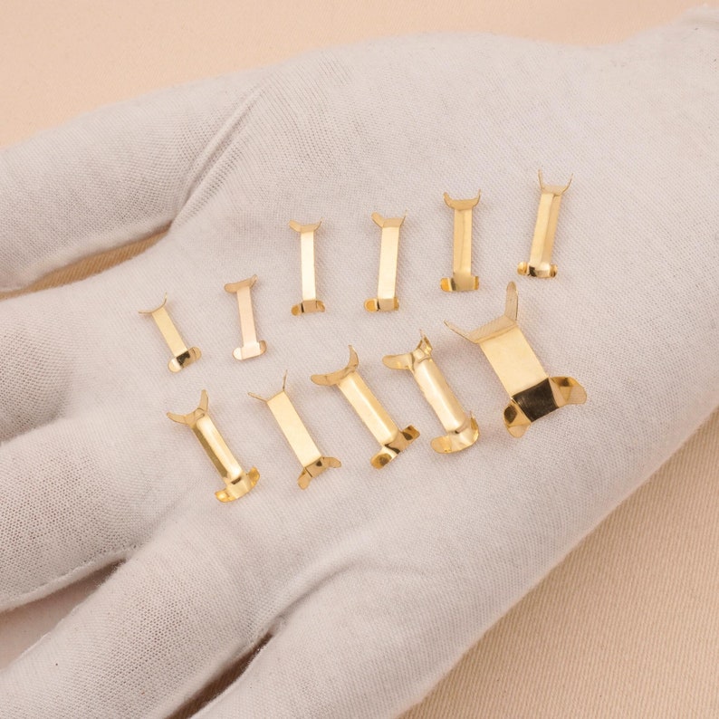 Puede incluir: Una colecci&oacute;n de peque&ntilde;os cierres met&aacute;licos dorados y brillantes. Los cierres son rectangulares con un dise&ntilde;o curvo en forma de ala en cada extremo. Est&aacute;n dispuestos sobre una superficie blanca.