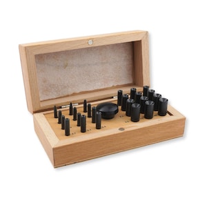 May include: A wooden storage box containing a set of black metal watch case press dies. The set includes various cylindrical tools of different diameters, each marked with numerical sizes, arranged in a light-colored wooden base with a hinged lid.