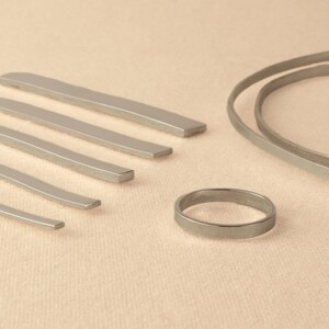 May include: A set of silver metal rings and strips of varying sizes. The rings and strips are arranged on a light beige surface.
