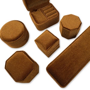 Könnte beinhalten: Fünf braune Samt-Ringboxen in verschiedenen Formen und Größen. Die Boxen sind alle aus einem weichen, strukturierten Stoff gefertigt und haben ein klassisches, elegantes Design.