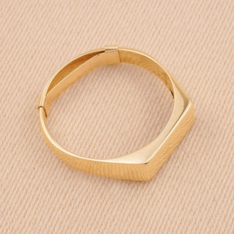 Puede incluir: Un brazalete dorado con un dise&ntilde;o geom&eacute;trico. El brazalete tiene forma rectangular en un lado y una banda curva que se conecta a un cierre. La joya est&aacute; hecha de un metal brillante y reflectante.