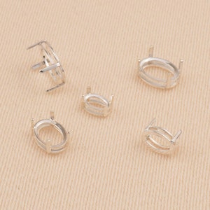 May include: Five silver-toned oval-shaped jewelry settings, each featuring a hollow center and four vertical prongs designed to hold a gemstone. The settings are arranged in a scattered pattern, showing various angles of the metal components.