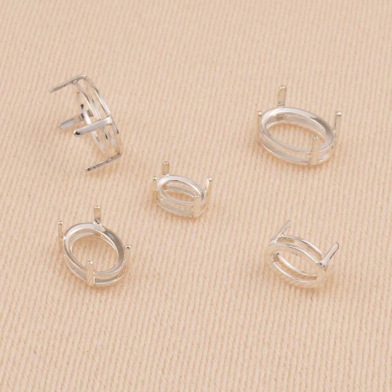 May include: Five silver-toned oval-shaped jewelry settings, each featuring a hollow center and four vertical prongs designed to hold a gemstone. The settings are arranged in a scattered pattern, showing various angles of the metal components.