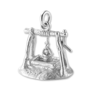 Puede incluir: Un charm plateado con forma de pozo. El pozo tiene un cubo colgando de una viga de madera, sostenido por dos postes. El charm está detallado con una base texturizada y un lazo para sujetarlo a una pulsera o collar.