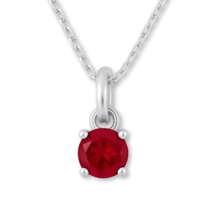 Puede incluir: Un collar de plata con un colgante de gema roja oscura y redonda. El colgante está sujeto por cuatro pequeñas garras y cuelga de una delicada cadena. La gema tiene una superficie facetada.