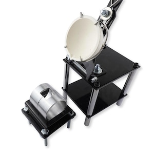 May include: A metal casting machine featuring a black frame with chrome support pillars, a white ceramic crucible held by a metal arm, and a separate cylindrical metal mold base. The apparatus includes various bolts, nuts, and mechanical joints.