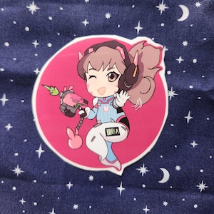 May include: A pink and white cartoon character with brown hair and pink bunny ears holding a green and black weapon. The character is wearing a blue and white jumpsuit with the word "DEKA" on the leg. The character is on a pink circle with a white outline.