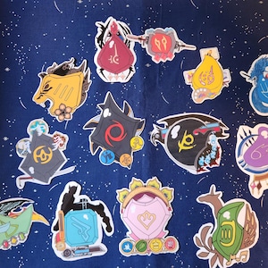 May include: A collection of 12 colorful stickers featuring various designs, including a purple creature, a green creature, a yellow creature, a red shield, a black shield, a blue shield, a yellow and black shield, a green and yellow shield, a pink shield, a black and yellow creature, a blue creature, and a green creature.