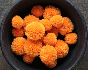 May include: A dark, round bowl filled with bright orange pom-poms. The pom-poms are fluffy and densely packed, creating a textured appearance. The bowl sits on a dark wooden surface, providing a contrasting backdrop.