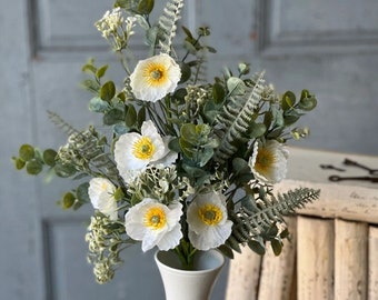May include: A white ceramic vase with a narrow neck and a wide base, filled with a bouquet of white flowers with yellow centers and green leaves.