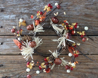 May include: A circular autumn wreath featuring miniature pumpkins, berries, and straw accents. The wreath is adorned with brown, orange, and red elements, including small pinecones and leaves. The wreath is set against a rustic wooden background.