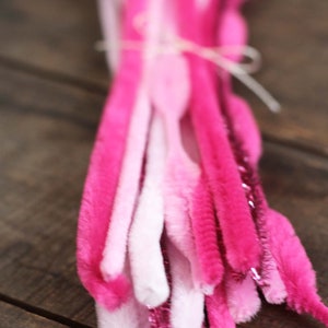 May include: A bundle of pink and white pipe cleaners tied together with a white string. The pipe cleaners vary in shades of pink, from light to dark, with some featuring a sparkly, glittery texture. The bundle is set against a wooden background.