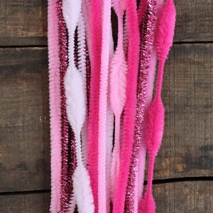 May include: A collection of pink and white pipe cleaners in varying shades, including hot pink, light pink, and white. Some pipe cleaners have a glittery, metallic finish. The arrangement is against a dark wood background.