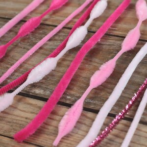 May include: An assortment of pink and white pipe cleaners arranged on a wooden surface. The pipe cleaners vary in shades of pink, from light to hot pink, and some have a fluffy, textured design. A few are white with a textured design.