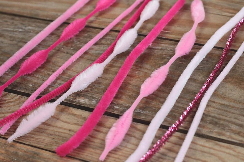 May include: An assortment of pink and white pipe cleaners arranged on a wooden surface. The pipe cleaners vary in shades of pink, from light to hot pink, and some have a fluffy, textured design. A few are white with a textured design.