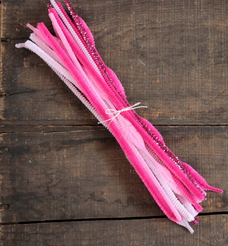 May include: A bundle of pink and white pipe cleaners tied with a white string. The pipe cleaners vary in shades of pink, from light to dark, with some featuring glitter.