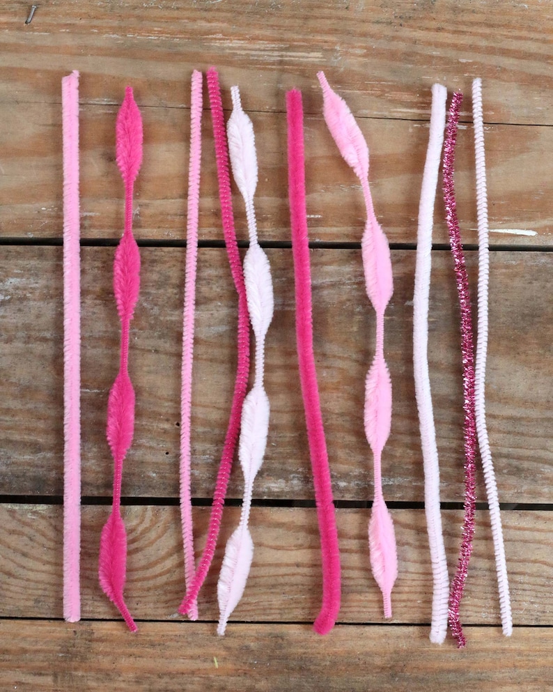 May include: An assortment of pink and white pipe cleaners and chenille stems. The stems vary in texture and design, including solid, twisted, and feather-shaped elements. The color palette ranges from light pink to deep fuchsia, with a touch of glitter.